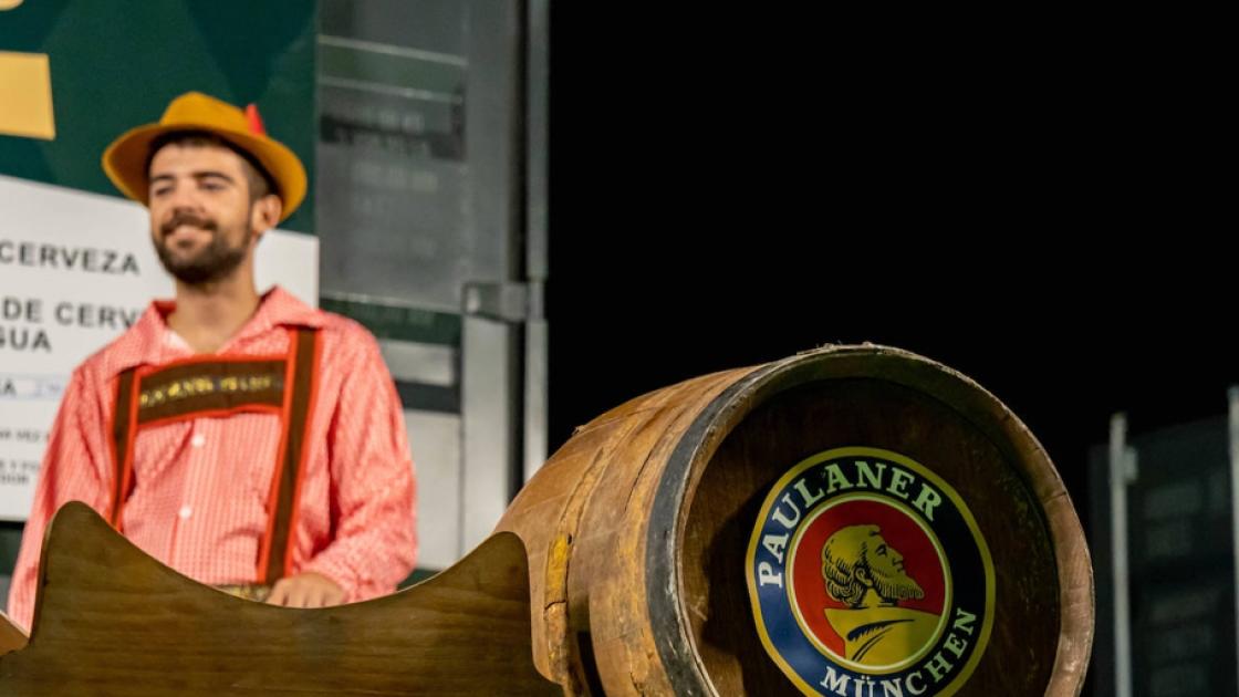 Un hombre vestido de manera típica frente a un barril de cerveza de madera con la marca Paulaner